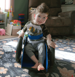 A little boy with brown hair sits in a blue wheelchair in his living room. There are toys behind him and sunlight is coming in the window. He is smiling at the camera.