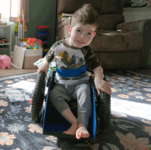 A little boy with brown hair sits in a blue wheelchair in his living room. There are toys behind him and sunlight is coming in the window. He is smiling at the camera.