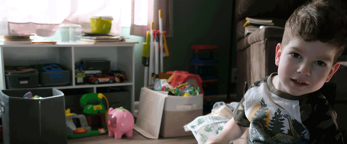 A little boy with brown hair sits in a blue wheelchair in his living room. There are toys behind him and sunlight is coming in the window.