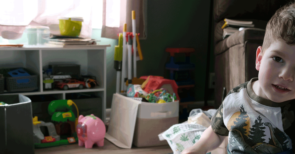 A little boy with brown hair sits in a blue wheelchair in his living room. There are toys behind him and sunlight is coming in the window.