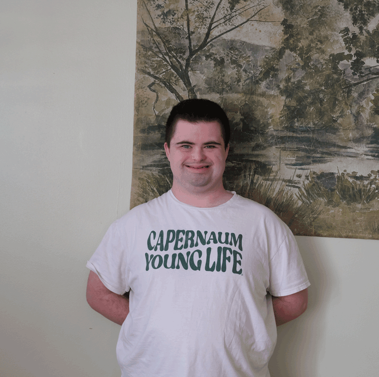 A photo of a young man with short dark hair standing in front of a green wall hanging.