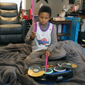 A little boy with dark skin and dark hair wearing a "SpaceJam" jersey plays the drums with pink sticks.