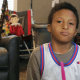 A little boy with dark skin and dark hair wearing a "SpaceJam" jersey holds pink drumsticks and looks at the camera.