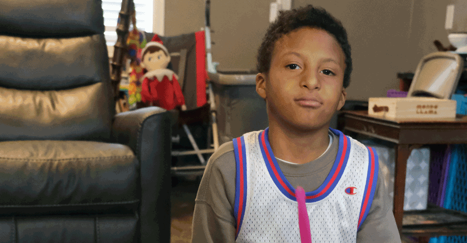 A little boy with dark skin and dark hair wearing a "SpaceJam" jersey holds pink drumsticks and looks at the camera.