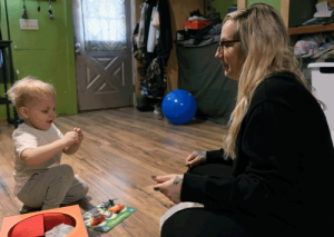 A little boy with blonde hair smiles while holding a plastic animal. He is sitting on the floor across from his mother.
