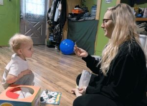 A little boy with blonde hair looks at his mother as she holds up a small plastic dinosaur.
