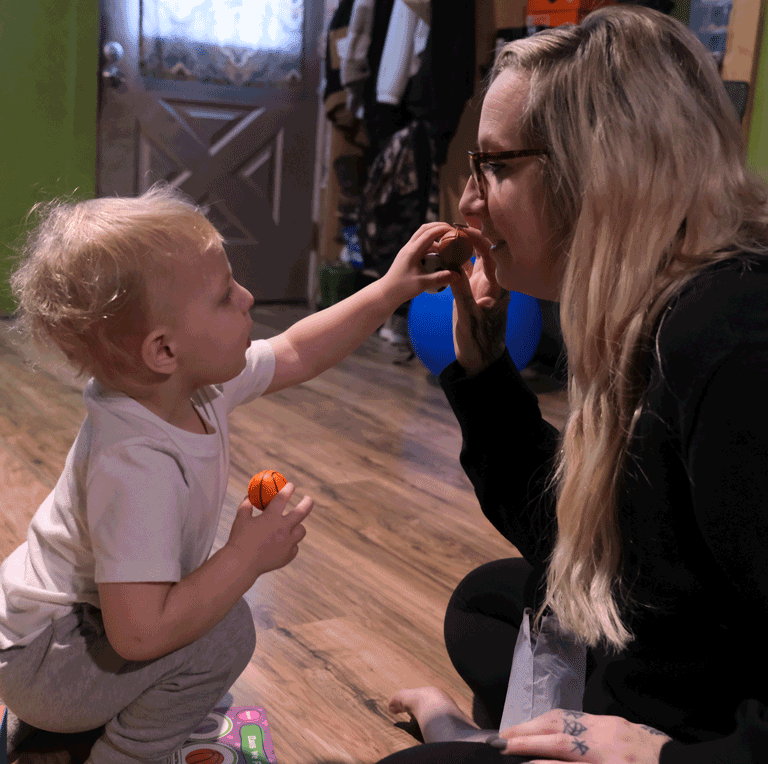 A little boy reaches for a tiny football held by his mother. He is holding a little basketball.