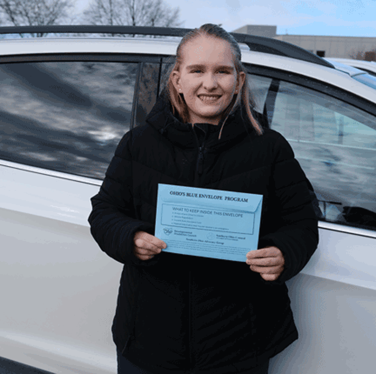 A close up image of a woman wearing a black coat stands in front of her car holding a blue envelope.