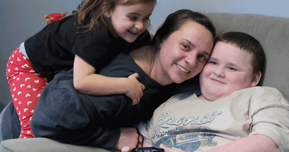 A boy sits in a chair and gets a hug from his mother and little sister.