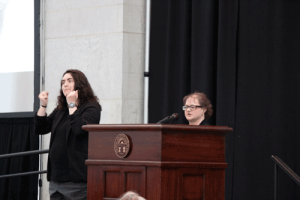 Hope Hindel, a woman with short brown hair, stands behind a podium at the the Ohio Statehouse. An ASL interpreter stands  next to her. 
