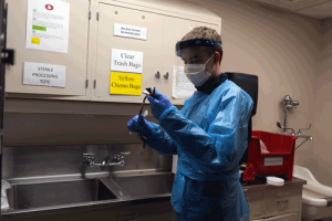 A young man is wearing a full body blue gown and a mask and face shield. He is cleaning lead wires.