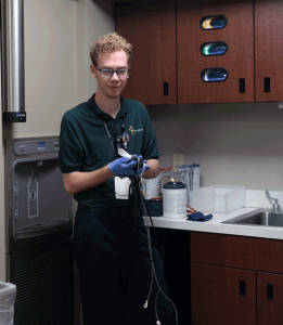A young man with short curly hair cleans a piece of medical equipment with a disinfecting wipe. He is wearing a green Project SEARCH polo shirt.