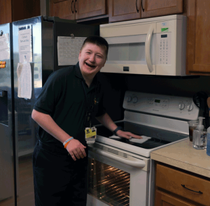A young man wearing a a green Project SEARCH polo shirt wipes off a stove. He has a big smile on his face.