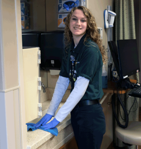 A young woman with curly hair cleans a counter with a blue cloth. She is wearing a green Project SEARCH polo shirt.