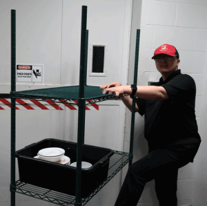 A young man poses for a photo with his dish cart. He is wearing a red and black uniform.