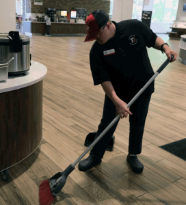 A young man sweeps the floor at the Curtis Hall Dining Hall. He is wearing a red and black uniform.