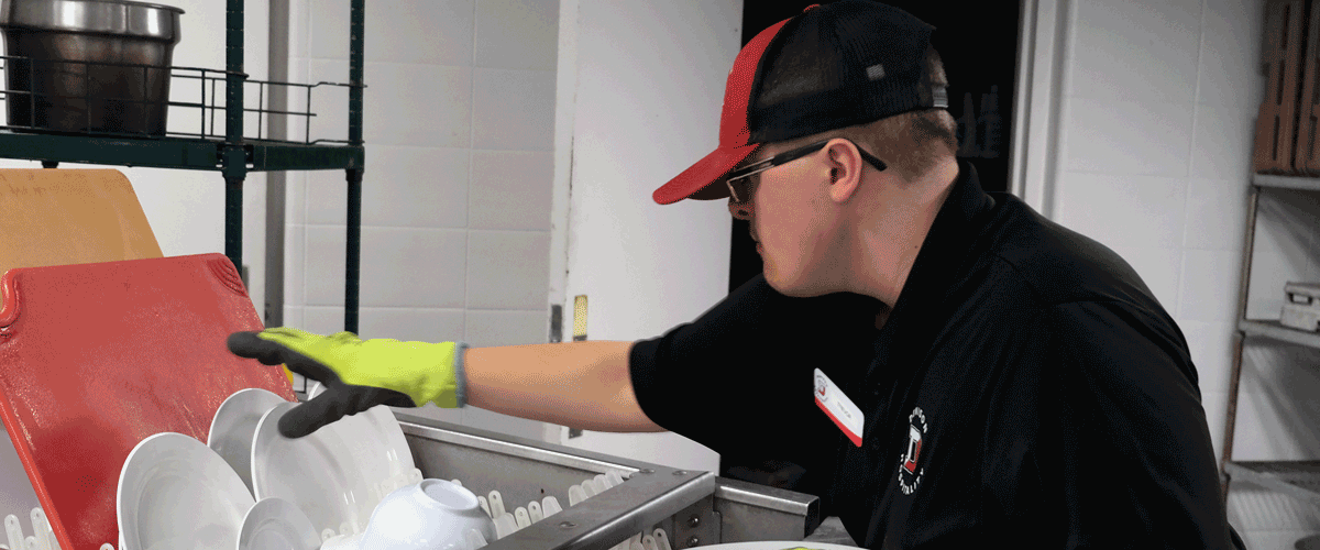 A photo of a young man unloading clean dishes from a dish machine. He is wearing a red and black uniform.