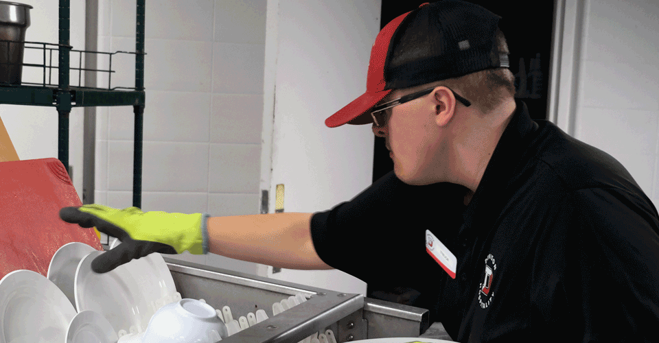 A photo of a young man unloading clean dishes from a dish machine. He is wearing a red and black uniform.