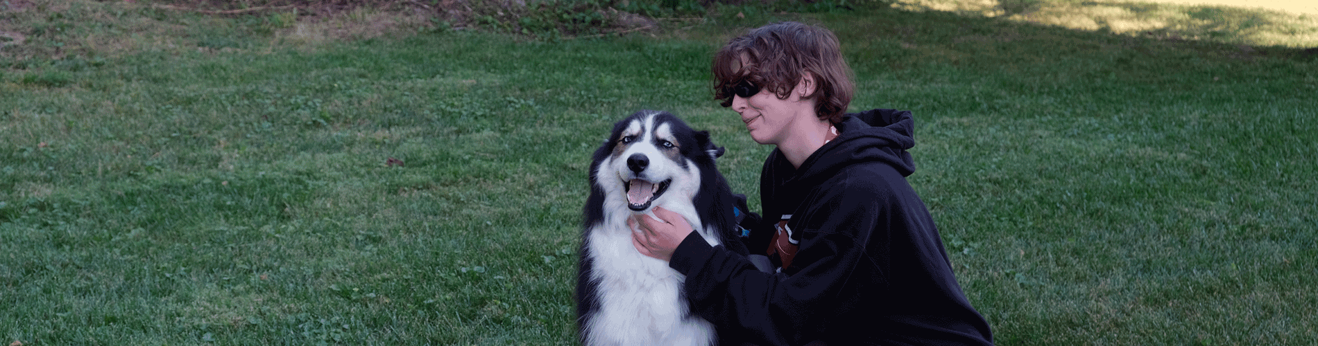 A young woman in a black hoodie kneel downs to pet her dog. The dog is a black and white dog with lots of fur. They are outside in the grass with a tree in the background.